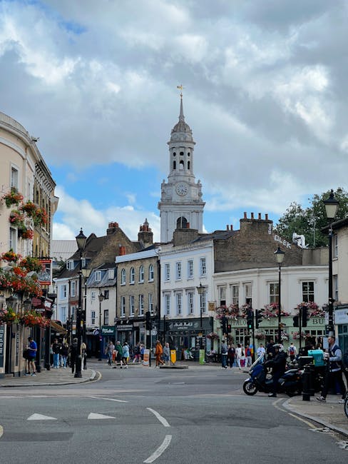A bustling street scene in Mill Hill NW7 featuring a traditional white clock tower with a gold weather vane at the top, surrounded by Victorian and Georgian terraced buildings with decorative window b