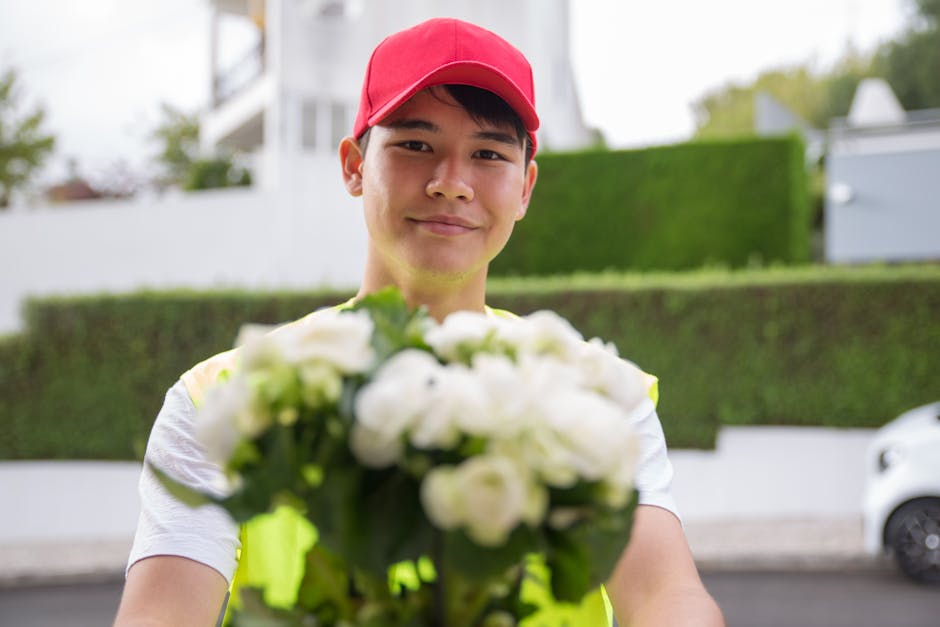 A delivery person wearing a red cap and a high-visibility yellow vest is standing outdoors on a residential street, holding a bouquet of fresh white roses and peonies. The bouquet is neatly arranged, 