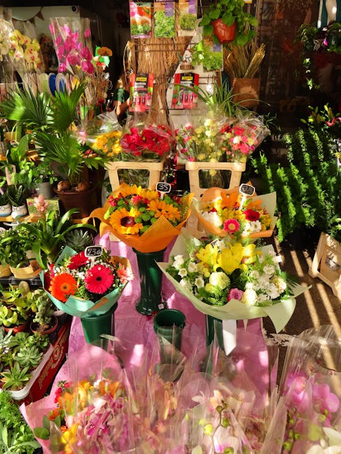 A vibrant display of fresh floral bouquets arranged on a pink table in a florist shop. The bouquets feature an assortment of bright flowers including yellow daisies, pink gerbera daisies, white chrysa