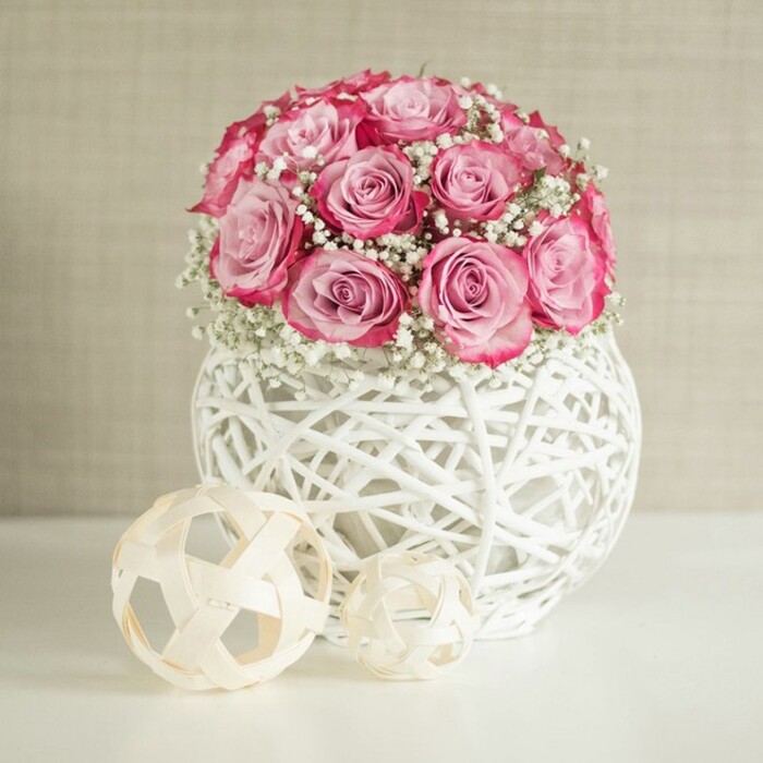This image portrays a serene tabletop arrangement that combines delicate florals with modern, textured decor, reminiscent of a carefully styled living room in Mill Hill. Dominating the composition is a lush, spherical bouquet of pink roses, the blooms gathered tightly together to create a full, domed shape that feels both intentional and generous. Each rose showcases a refined gradient of pink, beginning with a gentle pastel blush at the centre, then gradually deepening into a richer, more saturated pink towards the edges of the petals. This two-toned effect gives each flower a painterly quality, as if brushed with colour, and when viewed together, the roses form a soft yet vibrant focal point. Interspersed among them are numerous sprigs of white baby's breath, their tiny clustered blossoms creating a misty halo around the roses. The gypsophila introduces a cloud-like texture that lightens the arrangement, echoing the airy skies often seen over Arrandene Open Space. The bouquet is cradled in a unique spherical white vase crafted from intertwining strands of material that resemble white-painted wicker or rattan. This openwork design forms a basket-like lattice that feels both contemporary and slightly rustic, allowing glimpses of shadow and light to play across the smooth white surface beneath. To the left of the vase, two decorative woven spheres in pale beige rest casually, one larger, one smaller, fashioned from slim natural wood or fibre strips arranged in open, geometric tangles. They mirror the texture of the vase and introduce a soft, natural accent, as if chosen to complement a neutral-toned interior near Mill Hill Broadway. The background is a gently textured wall in warm beige or cream, softly illuminated by even, diffused light. Together, the arrangement and its setting convey a sense of tranquility, effortless elegance, and understated romance, perfect for marking a quiet anniversary dinner at home or brightening a calm reading corner.