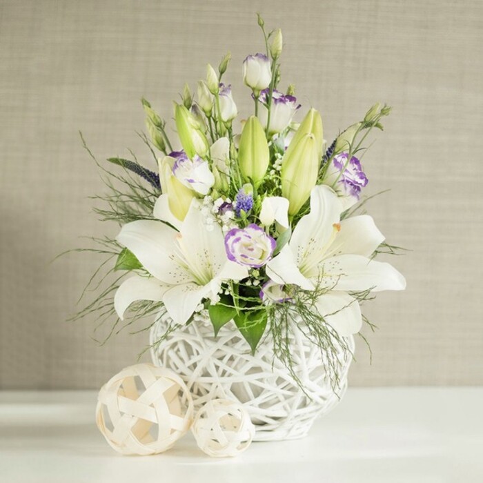 A tranquil floral centrepiece in shades of white, cream, and lilac is shown resting on a pure white surface, the kind you might see decorating a wedding table in a Mill Hill marquee. The arrangement is set in a round white vase with a striking open-weave, basket-like texture, its airy lattice catching the light and lending a stylish yet natural character. From this base, several white lilies extend upward, some blooms fully open with elegant, flared petals and clearly visible stamens, while others remain closed as elongated, pastel green buds, adding height and freshness.   

Interwoven among the lilies are delicate lisianthus flowers, their soft white petals edged or kissed with subtle lavender-purple tones. A mix of buds and blossoms provides a pleasing rhythm, with tightly curled buds giving way to layered, rose-like blooms. Tiny white filler flowers, reminiscent of baby's breath, are dusted throughout the display, and a few deeper violet accents hide between the stems, giving a touch of richness to the light colour palette. The arrangement is supported by generous greenery: feathery fronds, perhaps similar to asparagus fern or soft ruscus, mingle with broader leaves to provide both texture and volume. Several stems of foliage drape slightly over the rim of the vase, softening its silhouette and lending a natural, informal grace.   

In front of the vase sit two decorative spheres, crafted from a pale woven material that echoes the vase's open lattice. The background shows a light beige wall with subtle horizontal lines, as if covered in fine linen, gently illuminated so the whites glow warmly. The result is a peaceful and sophisticated display, fitting for weddings, moments of remembrance, or refined home décor across Mill Hill and neighbouring Green Belt streets.