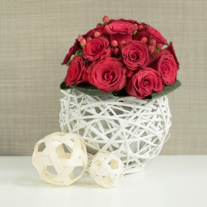 The photograph presents a romantic floral centerpiece that could grace any elegant wedding table in Mill Hill, its focus a dense, dome-shaped bouquet of luxurious red roses. The roses are arranged so closely together that they form a continuous, velvety surface of crimson, with each bloom showing its own personality. Some are still gently cupped, their petals layered tightly, while others have opened further, creating soft spirals and inviting shadows at their centres. The rich red tones are subtly varied, giving the bouquet a natural depth that feels both lush and refined. Nestled among the roses are clusters of red Hypericum berries, their smooth, glossy forms providing a playful counterpoint to the softness of the petals. Beneath the blooms, hints of dark green foliage peek out, adding structure and framing the arrangement with a quiet touch of natural contrast. The bouquet rises out of a distinctive spherical vase created from white-painted wicker or twigs. The branches are carefully woven into an open lattice that feels airy and modern, with enough negative space to let the eye wander through to the stems and the gentle interplay of light and shadow inside. This sculptural vase stands on a bright white tabletop, whose smooth, reflective surface mirrors a hint of the vase's round shape and the deep reds above. To the left, two decorative wicker balls in light, natural wood tones rest on the same surface. The larger sphere sits closer to the front of the frame, while the smaller one is positioned slightly behind, creating a pleasing visual echo of the vase's woven texture and rounded form. The backdrop is a softly focused expanse of neutral beige or taupe, reminiscent of the serene interiors of North London homes near Mill Hill Broadway and surrounding leafy streets. The entire composition gives off a sense of understated luxury and heartfelt romance, as if prepared by an experienced local florist for a wedding reception or intimate celebration.