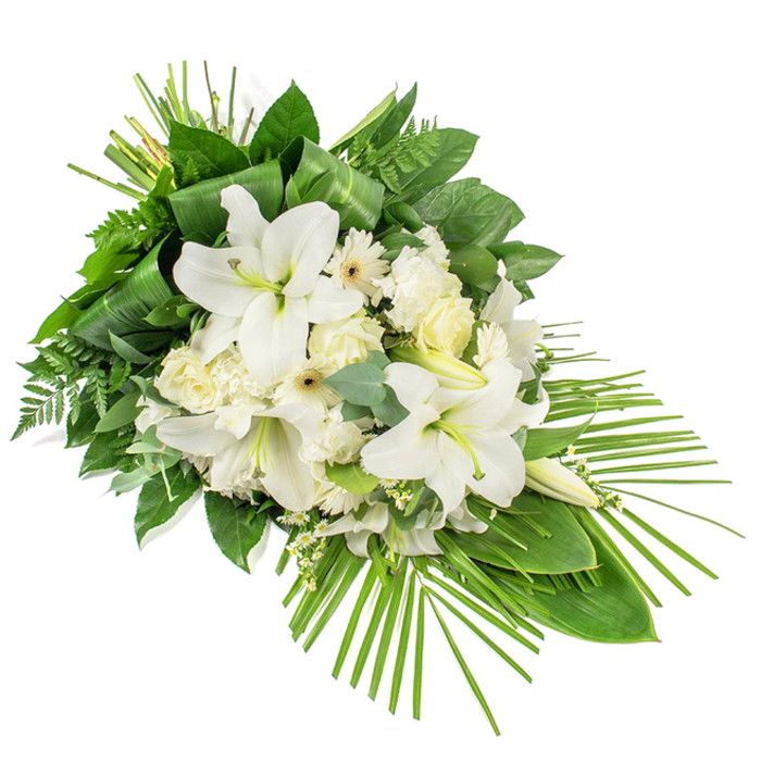 A striking, elongated funeral arrangement in pure white and deep green stretches diagonally across a white background, designed as a front-facing spray that tapers gracefully towards the lower right. At its heart, stately white lilies open into wide, star-like blooms, each petal smooth and slightly curved at the tips, exposing subtle green centres and softly powdered stamens. Several lily buds rise above, closed and sculptural, adding height and a sense of continuity. Encircling these lilies, groups of white and cream roses appear at different stages of opening, some tight and compact, others loosening into soft spirals that catch the light. Nestled between them, frilled white carnations provide volume and texture, their ruffled petals suggesting gentle movement. White gerbera-style flowers, with neat rings of petals and delicate cream or darker centres, are interspersed through the design, lending a fresh, modern feeling. Fine, airy white filler flowers weave among the main blooms, creating a light veil that softens the transitions between petals and leaves. The base of the spray is formed by a rich mix of greenery: broad, glossy dark leaves give weight and structure, feathery fern fronds soften the edges, and distinctive eucalyptus stems with round, blue-green leaves add coolness and a faint herbal impression. At the upper end, long, narrow leaves are folded and bound into upright bundles, adding a clean vertical accent. From the lower right, multiple slender palm fronds fan outward in a sweeping arc, emphasizing the teardrop shape and drawing the eye along its length. The arrangement feels composed and peaceful, a fitting tribute for a funeral or memorial gathering in Mill Hill, perhaps resting near a window overlooking local gardens or placed by the altar at a nearby church.