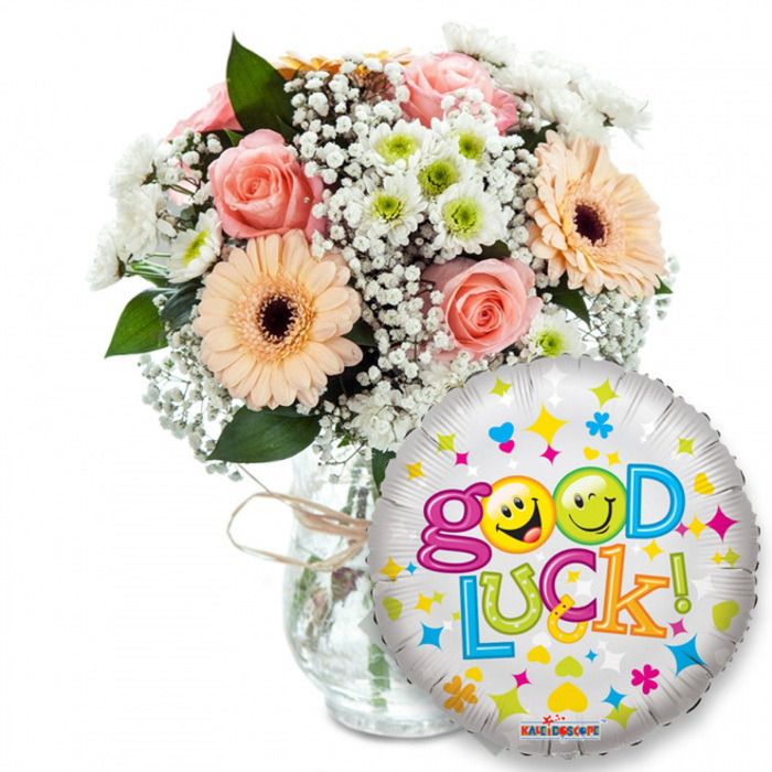 Set against a pristine white backdrop, this cheerful 'Good Luck' arrangement combines garden-fresh blooms and a playful foil balloon to create an immediate sense of celebration and encouragement. The bouquet, seated slightly left of centre in a clear glass vase tied with rustic twine, is crafted from pale peach and soft pink roses whose layered petals suggest a tender, floral perfume. Complementing them are peachy-orange Gerbera daisies with rich dark centres that add graphic warmth, while clouds of tiny white baby's breath (Gypsophila) and tight clusters of white-and-lime chrysanthemum florets provide delicate texture and bright contrast. Deep green foliage peeks through like punctuation, grounding the pastel palette and lending dimension when seen from the front or the side.

Prominently placed to the right and overlapping the vase, a shiny silver foil balloon catches the light and repeats the optimistic message: Good Luck! The balloon's metallic surface is decorated with colourful hearts, stars and confetti shapes in blues, pinks, yellows and greens; the word Good features two sunny yellow smiley faces in place of the 'o's-one cheekily winking, the other sticking its tongue out-while the rest of the letters appear in lively, contrasting hues. The small KALEIDOSCOPE logo is visible at the bottom rim of the balloon. As an arrangement prepared by Flowers Mill Hill, I imagine it being chosen for farewells, exam-day support or new beginnings and delivered with care to addresses across Mill Hill Broadway, Hale Lane and the Village, carrying both colour and thoughtful intent.