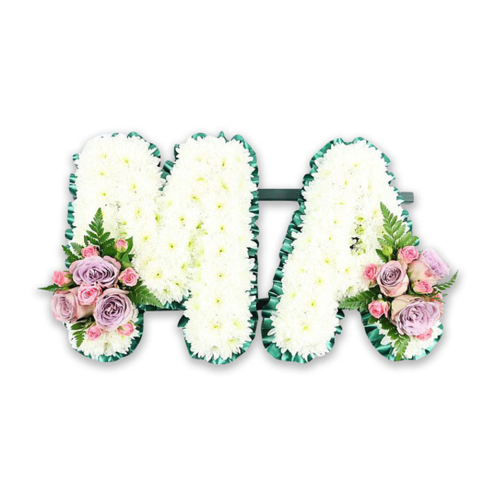 This personalised funeral tribute displays the letters MA in strong, clear capitals, a design often chosen to honour a mother, Mama, or dear matriarch in a family. The arrangement is photographed against a plain white background, allowing the details of the flowers and craftsmanship to take centre stage. Both letters are completely surfaced with small white chrysanthemums, tightly massed to form a smooth, cushioned layer of blooms. The petals are fine and lightly feathered, overlapping to create a plush, cloud-like texture that speaks of comfort and peace. Encircling the perimeter of each letter is a deep green trim, carefully applied to follow every contour. This border, likely formed from pleated satin ribbon or slim bands of foliage, provides a strong visual frame that sets off the purity of the white flowers. It also helps the letters read clearly in a funeral setting, whether placed near the coffin or displayed at a crematorium chapel serving Mill Hill and the surrounding North London area. At the base of the left stroke of the M and the right stroke of the A, tasteful flower clusters introduce soft colour and added depth. Within each cluster, pastel pink and lilac roses mingle with smaller pink spray roses, some blooms fully open while others remain partly closed, suggesting a gentle unfolding of memories. Fronds of fresh green fern emerge from beneath the roses, adding lightness and a natural, garden-like feel reminiscent of the greenery around local open spaces. The overall effect of the tribute is poised and heartfelt, bringing together purity, tenderness, and enduring affection in one simple yet deeply expressive floral piece.