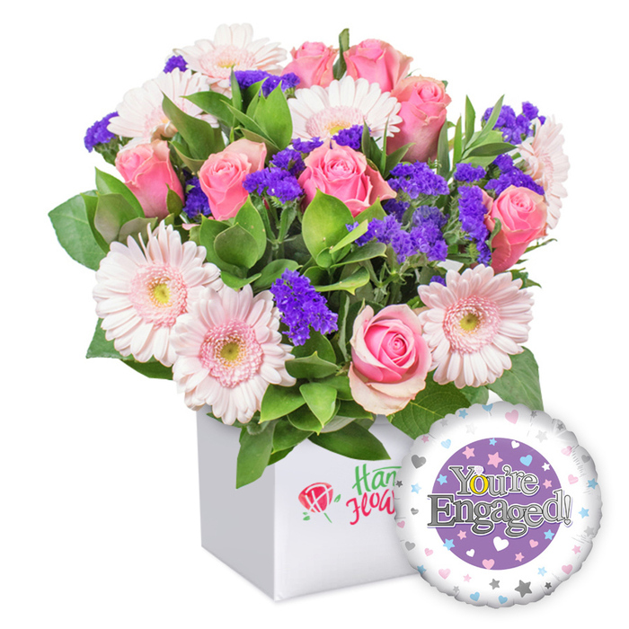 Elegantly arranged in a crisp white square box, this engagement bouquet radiates tender romance with contemporary flair, a signature offering for Mill Hill residents. The soft pink roses stand out, their petals unfurling in creamy spirals from tight buds to open blossoms, perfume light and sweet in the air. They mingle with pastel pink gerbera daisies, their white-edged petals haloed by pale yellow centers, creating a buoyant, sunlit mood. Interspersed among them are clusters of deep purple statice, lending depth and a whisper of mystery to the palette. A lush veil of green foliage weaves through the stems, providing a garden-fresh backdrop that evokes a quiet courtyard in Mill Hill East.
The blooms are neatly nestled in a clean, square white box, its front bearing a delicate red rose mark and green script that hints at local floristry craft without naming brands. In the lower right, a glossy foil balloon completes the gift: a round shape with a purple center bearing You're Engaged! in silvery type, and the golden 'O' styled as a ring. The outer white rim is dotted with pastel hearts and tiny silver stars, echoing the romance of the moment. This arrangement is a timeless congratulations for an engaged couple, available for dependable delivery around Mill Hill, Mill Hill East, and nearby streets, and well suited to same-day gifts that brighten a doorstep or office.
