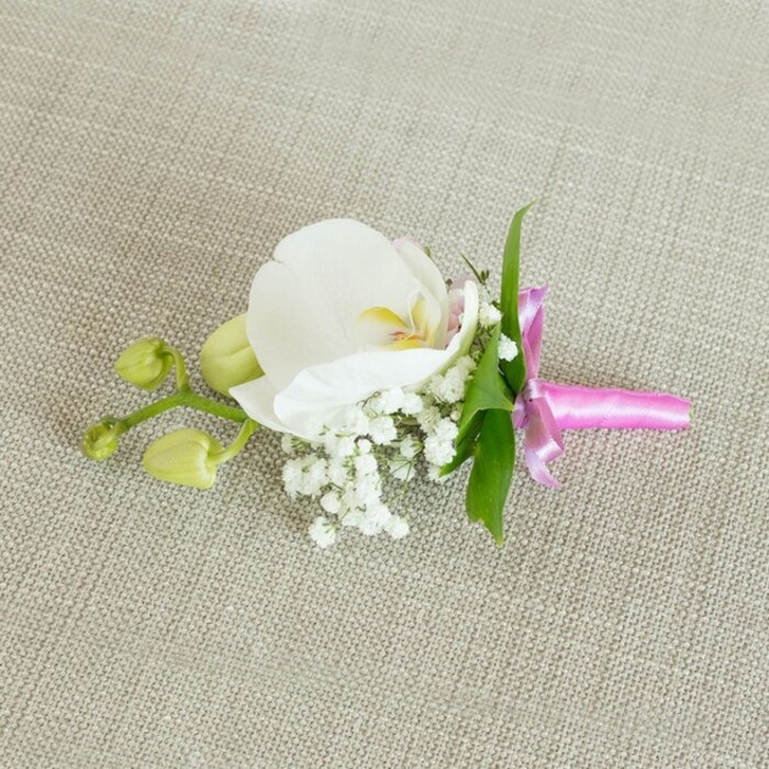 A timelessly elegant orchid boutonniere or corsage is photographed lying horizontally on a softly textured, oatmeal-coloured linen surface, the fabric's gentle weave catching the light in fine, understated lines. This neutral background, reminiscent of table linens set out in a Mill Hill family home before guests arrive, allows the delicacy of the flowers to stand out without distraction. At the centre of the piece, a single large white Phalaenopsis orchid blooms in full, its broad petals smooth and slightly arched, giving the impression of poised movement. The creamy white petals surround a small, intricate throat where subtle yellow and very pale pink details create a warm, inviting focal point. clustered coaxingly around the base of the orchid are numerous sprigs of white baby's breath. Each miniature flower dotting these sprigs adds to the cloud-like halo that wraps the main bloom in a soft, airy embrace, giving the arrangement a romantic, almost ethereal feel. From behind the flowers, slender, vivid green leaves stretch out lightly, bringing fresh colour and structure, and anchoring the design in natural foliage. To the left, a short branching stem bears a couple of tightly closed, pale green orchid buds. Their rounded, unopened forms suggest the promise of future blossoms, echoing the anticipation that surrounds weddings and formal gatherings across Mill Hill, from local churches to venues near Mill Hill Broadway. All the stems are carefully aligned and neatly secured with a vibrant fuchsia satin ribbon, wound smoothly around them and tied into a small, elegant bow. The bow's glossy sheen and strong pink hue offer a lively contrast to the soft whites and greens, injecting a modern, celebratory note into this otherwise classic design. Soft, even lighting falls across the composition, highlighting each petal edge, the tiny baby's breath flowers, the gentle curves of the buds, and the subtle grain of the linen, creating an impression of calm preparation just before a special occasion begins. This handcrafted accessory feels ideal for a groom's boutonniere, a groomsman's lapel, or a wrist corsage for an evening celebration, carrying with it the quiet sophistication and care of a well-planned event.