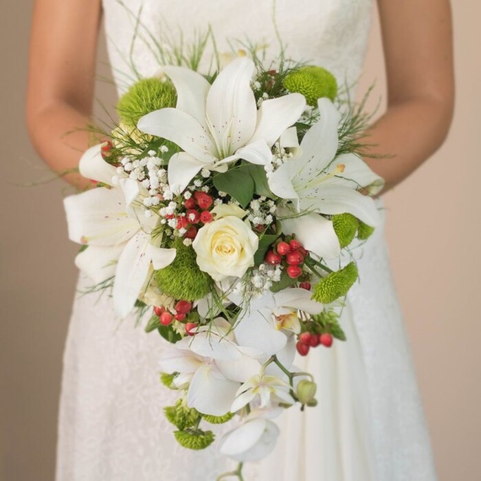 This close-up, taken just outside a Mill Hill venue, reveals an exquisitely crafted cascading bridal bouquet, assembled with a blend of tradition and modern flourish that defines Flowers Mill Hill's artistry. At its peak, dramatic white lilies with broad, trumpet-shaped petals stand tall and proud, each flower sporting delicate freckles of chestnut-red for visual intrigue. A lush cream rose tucked at the centre bestows a romantic, time-honoured charm, while spherical, lime-green cushion chrysanthemums leap out with cheer, calling to mind the vibrant greens of Mill Hill's open spaces. Glossy clutches of red hypericum berries are dispersed through the arrangement, their rich tones adding warmth and serving as celebratory nods to the neighbourhood's lively wedding culture. Cascading downward, white Phalaenopsis orchids lend an ethereal elegance, the shape of their delicate blooms creating a gentle flow that defines the bouquet's teardrop silhouette. Sprigs of white baby's breath and slender grass-like greenery bring ethereal softness, echoed by the rich green foliage providing structure and harmony. The faint lace pattern of the bride's gown is visible behind, setting an intimate, romantic backdrop for the vivid display. This bouquet-a showcase of bespoke North London style-mirrors the sense of community, natural beauty, and craftsmanship found throughout Mill Hill, making it an exceptional inspiration for local brides and wedding planners alike.