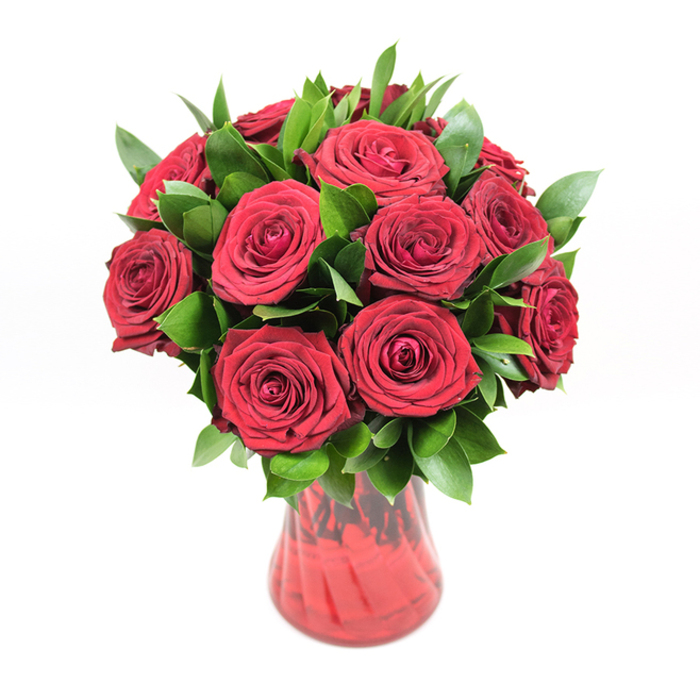 A dramatic, classic bouquet of a dozen or slightly more deep red roses is showcased in a richly tinted red glass vase, photographed against a flawless white backdrop that strips away distraction and focuses the eye entirely on the flowers. The roses themselves are at full, luxurious bloom, with velvety petals overlapping in tight, nest-like spirals that create a sense of depth and softness you can almost feel with your fingertips. Their colour is a striking crimson, closer to a ruby than a bright scarlet, and under the even studio lighting, each bloom reveals subtle gradients-almost black-red in the deepest folds and glowing, warm red along the outer curves. Interlaced with the blooms and spilling gently around the edges of the bouquet is a generous amount of lush dark green foliage, likely ruscus or similar hardy greenery. The leaves are smooth, pointed, and slightly glossy, catching glints of light that make them look freshly cut, as if they've just been conditioned in a Mill Hill flower shop before arranging. They provide a rich, supportive frame that accentuates the roses' intensity, giving the entire design a rounded, balanced silhouette. The vase is made of translucent red glass, with a softly curving body that narrows near the base and widens into a flared neck. A delicate ribbed or swirled texture runs through the glass, so that when the light hits, it seems to ripple with darker and lighter shades of red. Though the stems are partly obscured by the glass's rich colour, you can sense their straight, sturdy lines anchoring the design. This image evokes a carefully crafted romantic gift, the sort of bouquet someone in Mill Hill might have delivered from Flowers Mill Hill to a loved one near Mill Hill Park or along Bittacy Hill to mark an anniversary, Valentine's Day, or a heartfelt I love you without a single spoken word.