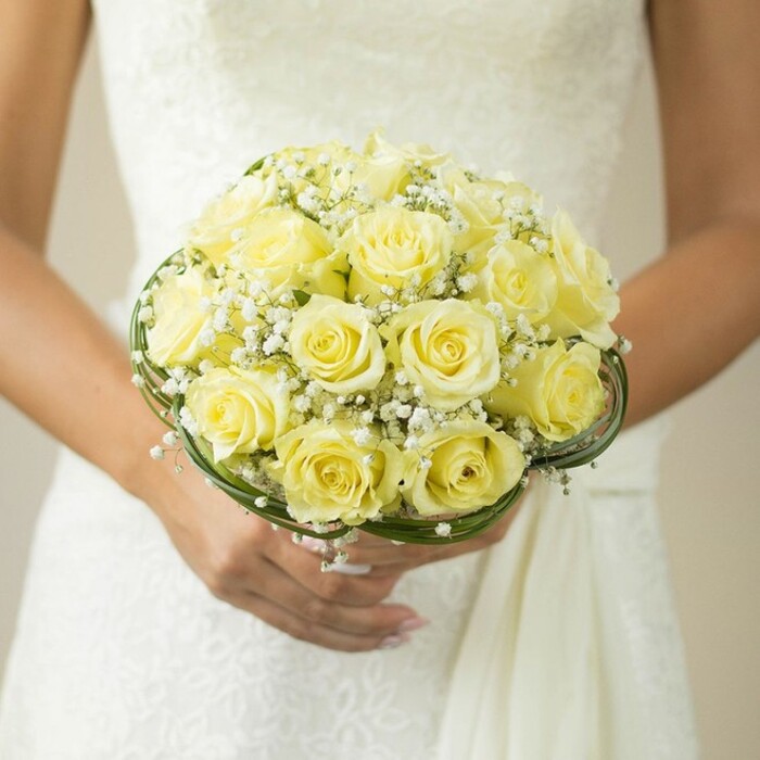 A timeless portrait from Mill Hill captures the pure elegance of a bride's bouquet held lovingly at mid-chest level, her hands gently cupping a lush round gathering of soft yellow roses. Every rose is in its prime, petals softly unfurled and pressed close together, forming an inviting, domed accent that glows with the gentle warmth of early sunshine over Mill Hill's gardens. Interspersed are wispy sprigs of baby's breath-tiny white blossoms that lend an airy lightness, like a cloud drifting above the rolling green spaces of North London. The bouquet is framed at its base by neat, looping lines of rich green-either slender foliage or meticulously wrapped wire-serving as an elegant border that echoes the decorative hedges along Daws Lane. The bride's perfectly manicured nails and simple wedding ring glimmer softly beneath the arrangement, while her wedding dress, with its textured lace or embroidery, creates a tranquil backdrop subtly blurred to draw focus to the floral artistry. This scene celebrates Mill Hill's tradition of thoughtful, beautiful occasions, offering a vision of sophisticated romance and fresh beginnings created with the skilled hands of Flowers Mill Hill's local team.