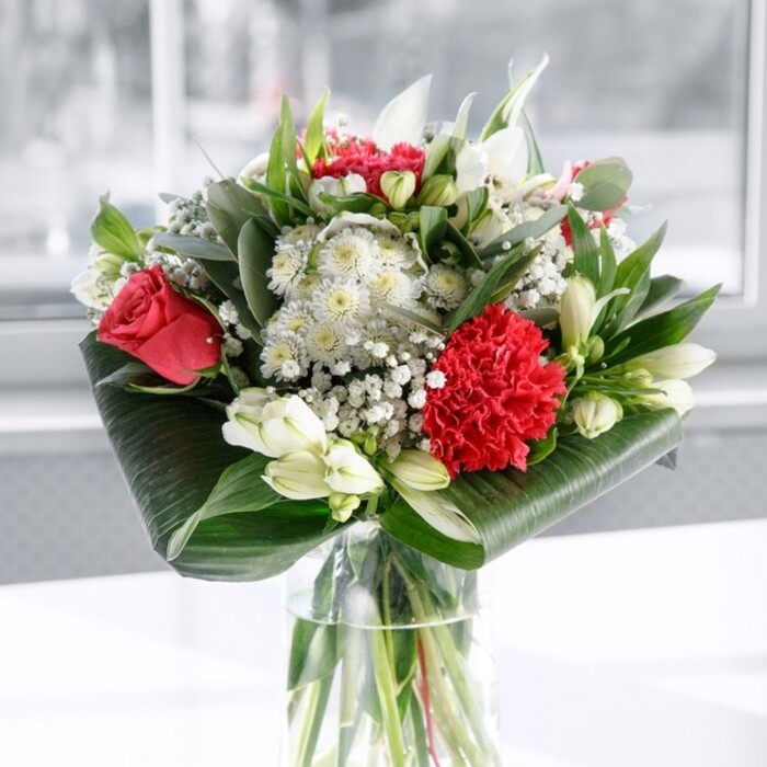 A richly romantic bouquet in tones of deep red, crisp white, and fresh green stands tall in a clear cylindrical glass vase, placed on a spotless white surface with a hazy, light-filled backdrop. The composition feels like a carefully arranged still life set in a Mill Hill townhouse, where natural daylight gently brushes each bloom. At the centre, several velvety red roses unfold just enough to show the spiral of inner petals, giving the piece its passionate heart. Nestled close by, a large, intricately ruffled crimson carnation forms a textured focal point, its frills creating shadows and highlights that draw the eye. Interspersed among these vibrant reds are graceful white flowers: slim-petalled alstroemerias lightly speckled inside, rounded chrysanthemum-style blooms, and a few plump white lily buds standing upright, not yet open but promising scent and drama in the days to come. Delicate sprays of white gypsophila, or baby's breath, thread through the bouquet like fine lace, lending an airy quality that softens the stronger lines. The base of the arrangement is elegantly framed by broad, dark green tropical leaves, wrapped and curled around the stems to form a polished, almost architectural collar. Mixed foliage, including eucalyptus-like sprigs and pointed, fresh green leaves, weaves between the blooms, adding depth and different shades of green. Through the vase you can see the neatly cut stems submerged in clear water, signaling freshness and the professional care typical of Flowers Mill Hill for local deliveries around Mill Hill East and the Broadway. The overall impression is uplifting and composed, perfect as a centrepiece for an intimate celebration, a thoughtful anniversary surprise, or a refined gesture of appreciation in any Mill Hill home or office.