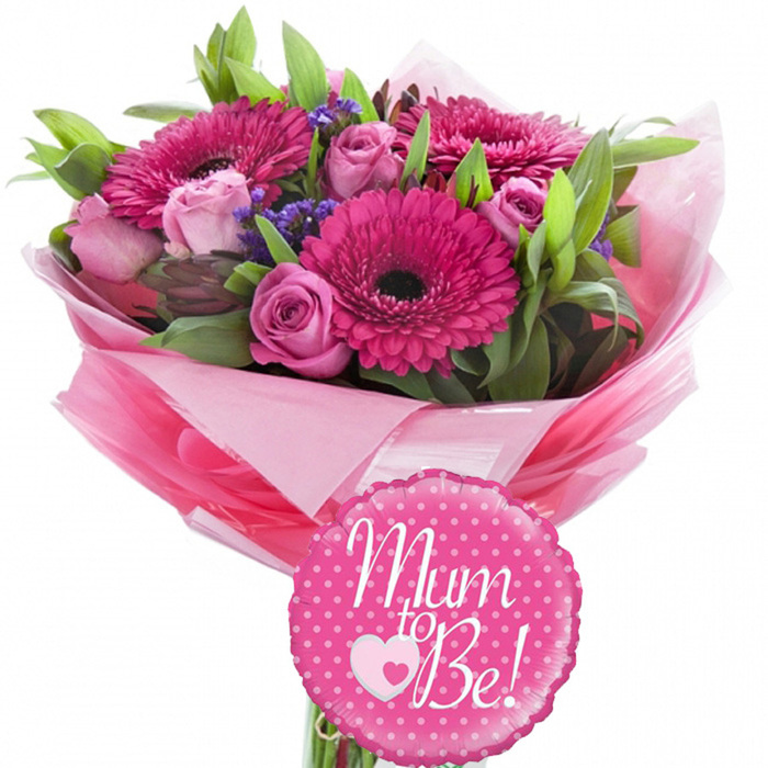 A tender yet vibrant floral gift designed to honour an expectant mother is shown against a clean white background, echoing the calm light one might find in a Mill Hill home on a bright morning. The bouquet forms a gently domed shape dominated by large fuchsia gerbera daisies, each with neatly layered petals encircling a deep, dark centre that gives the flowers an almost graphic, modern quality. Between these bold focal blooms, you can imagine the softness of delicate pink roses, some still held in tight buds and others starting to open, revealing their spiralling, velvety petals in shades of blush and pastel rose. Here and there, clusters of small, purple filler flowers emerge from the greenery, adding a textured, slightly wilder note that prevents the arrangement from feeling too formal and evokes the hedgerows and garden borders common in the Mill Hill area. A lush collar of mixed foliage, combining sleek, linear leaves with broader, glossy greens, frames the blossoms and helps the colours stand out, enhancing the sense of freshness you would expect from a bouquet prepared for same-day delivery. The stems are wrapped at the base in several layers of pink tissue paper; deep, vibrant pinks sit alongside softer, powdery tones, with an outer layer of clear cellophane creating a polished finish that rustles faintly when handled. Positioned prominently in front of the stems is a bright fuchsia foil balloon, round with a soft scalloped edge and decorated with an even pattern of white polka dots. At its centre, the words Mum to Be! appear in elegant white script, with a small white heart outline encasing a pink heart nestled between the words, a simple yet effective symbol of the love surrounding a new baby. The combined bouquet and balloon convey joy, affection, and anticipation, ideal for baby showers, hospital visits, or surprise deliveries to expectant parents living in and around Mill Hill Broadway or heading out towards Edgware.