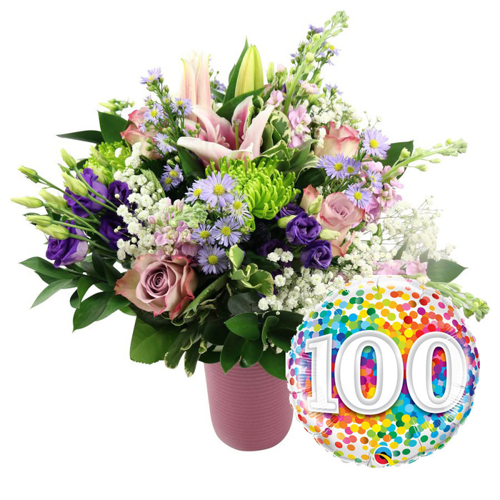 Framed by a spotless white background, this scene showcases a refined yet exuberant floral tribute to a 100-year milestone, as though ready to be carried through the quiet residential streets of Mill Hill East. A modern matte pink vase, gently ribbed and cylindrical, anchors the left side of the composition. From its narrow opening emerges a generous and deliberately sculpted bouquet, brimming with colour and texture. The upper section is dominated by elegant pale pink lilies, their petals sweeping outward in wide, starry forms, with a few buds rising just above the main cluster, promising even more blossoms over the coming days. Surrounding these focal blooms is a rich tapestry of roses in harmonious hues-blush, mauve, and dusty rose-each head fully formed and layered, contributing a sense of opulence and care. Threaded through the roses, velvety purple lisianthus blooms add depth, their gently ruffled edges catching imaginary light, while small lavender asters with bright yellow centres bring a refreshing field-flower charm. Scattered between the pastels, lime green button chrysanthemums appear as compact pom-poms, their bold colour enlivening the whole arrangement in the way spring grass brightens Mill Hill's open spaces. Airy white baby's breath meanders throughout, softening the transitions between blooms and imparting a light, misty texture. Various dark green leaves-some broad and glossy, others slim and more feathery-surround the flowers, giving structure and a natural finish. To the lower right of the bouquet, a round foil balloon glints cheerfully. Its surface is covered with dense, colourful confetti dots in red, orange, yellow, green, blue, and purple, while at the centre, the large white digits 100 stand proudly with a faint 3D shadow. This harmonious pairing of flowers and balloon captures the essence of a centennial birthday or anniversary, making it a fitting visual for a special Mill Hill delivery marking a century of life or love.