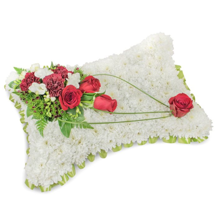 A beautifully constructed memorial pillow occupies the frame, every inch of its rectangular silhouette covered with hundreds of small, immaculate white chrysanthemum blooms to produce a dense, velvety texture that radiates serenity. Along the slightly raised border a ribbon of pale green or a thin band of foliage offers a soft hint of colour and a neat, finished edge. The principal floral accent sits at the upper left: several deep red roses - some tightly furled, others opening to reveal layered petals - are grouped with deep burgundy carnations whose ruffled edges add shadow and richness. Intermingled tiny white freesias or spray chrysanthemums give delicate highlights and a faint, clean fragrance that feels restrained and respectful. A lush array of green foliage, including slender fern fronds and broader glossy leaves, frames the cluster and deepens the contrast with the white chrysanthemum base. From the top-left grouping a trio of long, graceful green bear-grass stems sweeps diagonally across the pillow's surface toward the lower right, creating an elegant, unifying movement. This sweeping line appears to cradle a solitary red rose placed near the bottom right corner, a single, focused emblem of love and remembrance. The image's soft, natural light suggests an overcast morning in Mill Hill Village, and the overall composition conveys the considered skill of a local florist crafting a timeless funeral tribute intended to bring solace and honour a cherished memory.