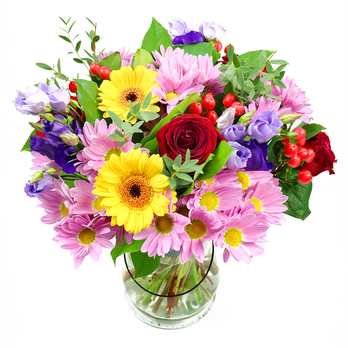 A rich, hand-crafted bouquet fills a clear glass vase, the arrangement standing alone against a white background so the viewer can focus entirely on the interplay of colour, texture, and form. Deep red roses form the heart of the design, their petals velvety and tightly layered, creating a sense of depth and timeless sentiment. Flanking them, two bold yellow gerbera daisies bring a cheerful brightness; their round outlines and brown-orange centres feel like little suns, reminiscent of clear afternoons spent around Mill Hill Park. Around these lead flowers, plush clusters of soft pink daisies or chrysanthemums extend the bouquet outward. Each bloom bears numerous fine petals radiating from a small yellow centre, producing a gentle, cottony effect that softens the stronger red and yellow tones. Emerging from between these pastel cushions are stems of purple and lavender lisianthus, their bell-shaped forms and subtly ruffled edges lending a cool sophistication that gives the bouquet a contemporary Mill Hill flair. Dotting the arrangement are small clusters of glossy red hypericum berries, adding playful texture and a festive accent, as if tiny ornaments have been tucked among the petals. The foliage is varied and fresh: broad, dark green leaves provide structure and contrast, while more delicate, light-green sprigs lift the composition and add a sense of natural movement. Through the glass vase, the evenly cut stems are clearly visible in clean water, evidence of careful preparation and attention to detail. This bouquet carries an atmosphere of joy and generosity, perfectly suited for birthdays, thank-you gifts, or a same-day delivery from Flowers Mill Hill to brighten a dining table or hallway console in a North London home.