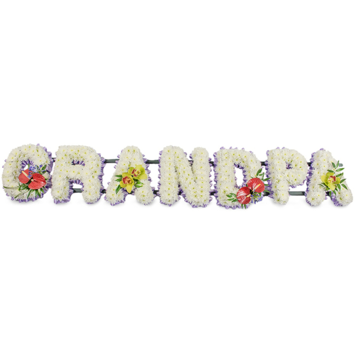 A striking, personalised funeral tribute dominates the image, spelling out GRANDPA in a continuous row of bold, three-dimensional letters made entirely from fresh flowers. Set against a simple white background, the design is easy to imagine in detail. Each letter is packed tightly with small white chrysanthemums, their neat, rounded petals creating a dense, plush surface that looks almost like a soft pillow of petals. This uninterrupted field of white conveys purity, peace, and a gentle, reflective mood, making it ideal for a memorial or funeral. Around the base of the letters, a slim edging of pale lavender ribbon forms a cool lilac halo, giving clear definition to the shapes and introducing a soft, calming tone. A sturdy green frame runs behind the letters, linking them together and ensuring the tribute can be displayed securely at a funeral service in Mill Hill, perhaps at a chapel used by local families from around Bunns Lane or Daws Lane. Thoughtful bursts of colour break up the white and add emotional nuance. The G on the left and the D towards the centre each feature clusters of deep red anthuriums, their glossy, heart-shaped petals and prominent yellow spadices nestled among dark green foliage and tiny purple filler flowers, suggesting steadfast love and heartfelt remembrance. The first and final A are embellished with yellow-green orchids, their graceful, arching petals lending a refined, elegant character to the piece. Crafted with care and local expertise, this tribute could easily be imagined as the work of Flowers Mill Hill, chosen by a family wanting a dignified, personal way to honour a beloved grandfather at a funeral, cremation, or graveside farewell.