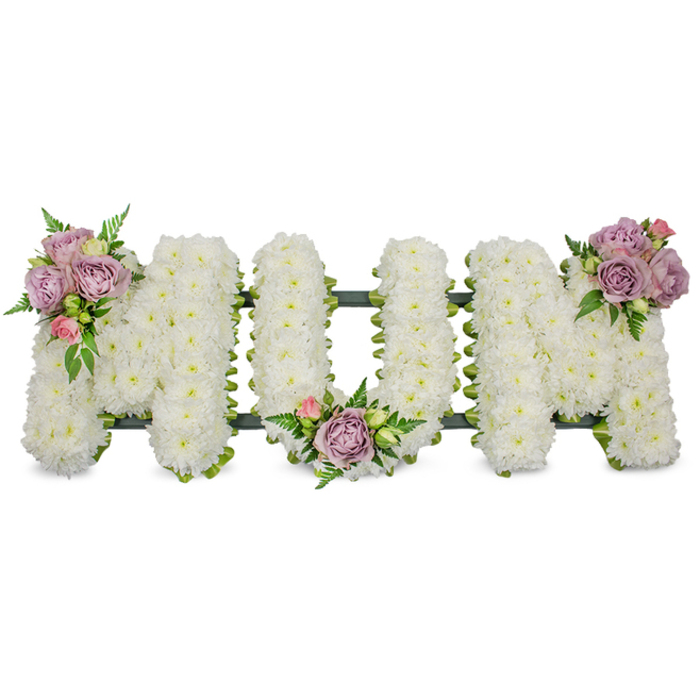 A pristine, three-dimensional floral memorial thoughtfully spells the word 'MUM' in bold capital letters, each letter artfully covered with hundreds of small, snowy-white chrysanthemums that create a consistently soft, textured surface. The white mass reads as a serene blanket, evoking peace and quiet dignity, while three clusters of subtle colour interrupt the whiteness with tenderness: lilac-lavender roses paired with delicate pale pink rosebuds sit on the upper left arm of the first 'M', a mirrored cluster nestles in the inward curve of the 'U', and a final grouping accents the upper right of the closing 'M'. Bright green leaves and wisps of fern thread through the clusters, giving contrast, movement and the visual impression of respiration within the form. A dark green structural trim - likely moss or ribbon over supporting board - peeks under the blooms and ties the letters together with a discreet, stabilising line.
Prepared by Flowers Mill Hill with the considered hand of a local florist who knows the pace of Mill Hill Village and the needs of families near The Ridgeway, this tribute reads as both personal and traditional. The arrangement sits well in a chapel or church hall bathed in soft morning light, and its gentle palette and careful craftsmanship make it an apt choice for funeral services, memorial gatherings, or graveside farewells. The overall effect is reassuring: an elegant, tactile memory piece that honours a mother with quiet beauty and local care.