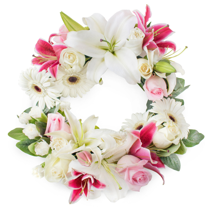 This open circular wreath, photographed against a pristine white background, forms a complete ring of flowers in harmonious shades of white, blush pink, and deep magenta, conveying both softness and strength. Dominating the composition are large white lilies, fully open and almost sculptural, their broad petals smooth and luminous, revealing delicate green centres and golden-tipped stamens that serve as subtle focal points. Around them, vibrant fuchsia and deep pink lilies weave through the design, their star-shaped blooms lightly freckled and slightly ruffled at the edges, adding a dynamic pop of colour reminiscent of bright florals seen in front gardens around Mill Hill. Between these larger elements, crisp white Gerbera daisies stand out with their bold, dark centres encircled by neatly layered petals, introducing a cheerful yet respectful contrast. The wreath is further enriched by soft pink roses-some tightly furled, others gently opening-symbolising affection and gentle remembrance, while creamy white roses in fuller bloom bring a timeless sense of grace. Smaller white blossoms, perhaps miniature carnations or sprays of petite roses, are carefully placed to fill gaps and build a dense, plush texture, ensuring the circular form feels complete and secure. A base of lush green foliage frames the entire wreath, with glossy leaves and hints of new growth adding depth and a natural, garden-inspired feel, much like a peaceful corner of Mill Hill Park. The overall effect is of a thoughtfully crafted funeral wreath, ideal for services in local churches or crematoriums, offering a tender expression of sympathy and a dignified way to honour a life well lived.