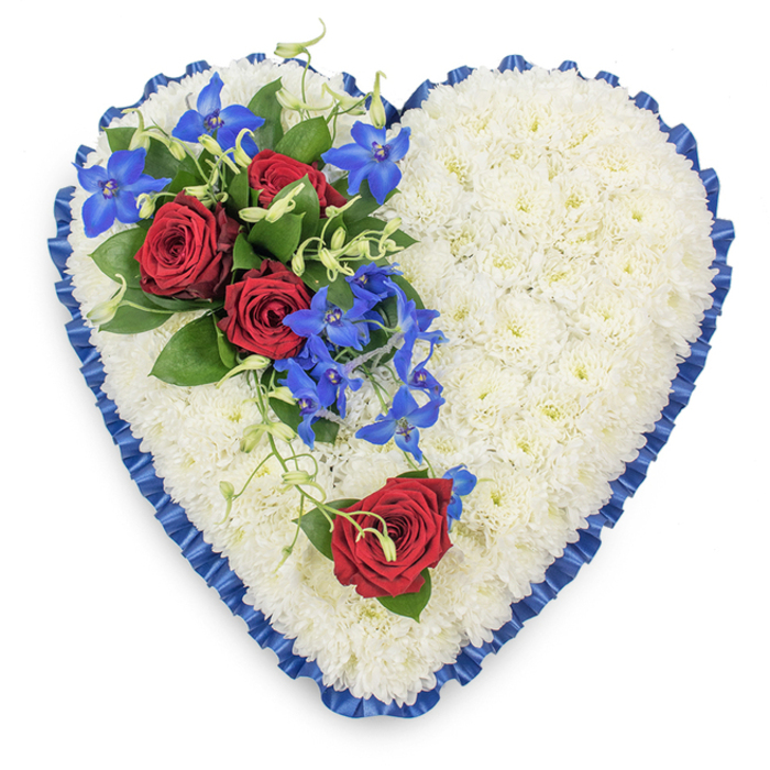 In the image, a refined heart-shaped funeral tribute is displayed in a clean, top-down view, ensuring that the entire design and its delicate details are clearly visible. The foundation of the heart is made from a tightly massed bed of white chrysanthemums, the small, daisy-like heads pressed closely together to form a continuous, cushion-like surface. Each flower's many petals create a layered texture, giving the heart a plush, almost cloud-like character that suggests peace and purity. Around this white expanse, a royal blue satin ribbon has been carefully pleated and gently ruffled, forming a decorative edging that follows the curves of the heart. The satin's soft sheen picks up subtle reflections, and its deep blue tone adds a note of quiet formality, much in keeping with the mood of a dignified service in Mill Hill. At the upper left portion of the heart, a vivid floral grouping serves as the focal point. Three deep red roses are fully open, their rich, velvety petals unfurling in full spirals, cradled by dark, glossy green leaves. They are accompanied by electric blue dendrobium orchids, whose elongated petals and slender stems give a light, cascading movement, almost as if they are gently flowing down the heart's surface. Fine green tendrils and small touches of foliage thread through the spray, softening its outline and drawing the colours into the surrounding white mums. On the lower right curve of the heart, a single, perfectly formed red rose is placed with intentional simplicity, mirroring the larger spray and offering a quiet point of reflection. Together, these elements create a tribute that feels balanced and deeply heartfelt, the sort of arrangement a local Mill Hill florist would prepare with care for a family seeking a graceful way to say goodbye and celebrate cherished memories.