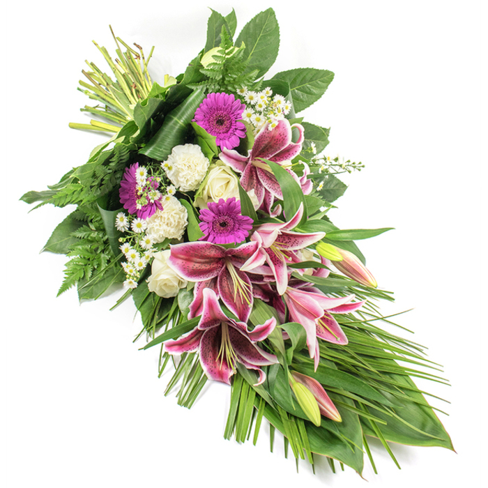 A beautifully composed funeral sheaf is shown on a white background, its elongated diagonal design creating a sense of calm movement from the top left to the bottom right of the frame. The stems are tightly bound at the upper left, then expand into a rich, layered cluster of flowers before softening into a trailing cascade of leaves and fronds. Dominating the central area are large, fragrant Stargazer lilies, their petals painted in vivid magenta and fuchsia with clean white margins and delicate burgundy speckles that draw attention to their curved, trumpet-like form. Some of the lilies are fully open, displaying their stamens and pollen, while others remain in elegant bud, tipped with hints of pink and green. Around them, full-headed magenta gerbera daisies add a modern splash of colour, offset by the gentle presence of creamy white roses and ruffled white carnations that convey innocence, reverence, and quiet affection. Fine sprays of small white daisy-type filler flowers thread through the arrangement, softening the borders between each bloom. All of this is framed by a generous collar of greenery: broad, dark, glossy leaves form a solid base, airy fern fronds reach outward with delicate fingers, and long, grass-like leaves extend and fall, echoing the line of the spray to enhance its graceful teardrop shape. The image captures the bouquet in clear, even light, making every texture-from velvet petals to smooth leaves-easy to imagine. It feels like a thoughtful tribute created by a local Mill Hill florist familiar with services at nearby churches along Mill Hill Broadway or Mill Hill East, ready to be delivered as a sincere expression of sympathy and remembrance for families across this quiet North London community.