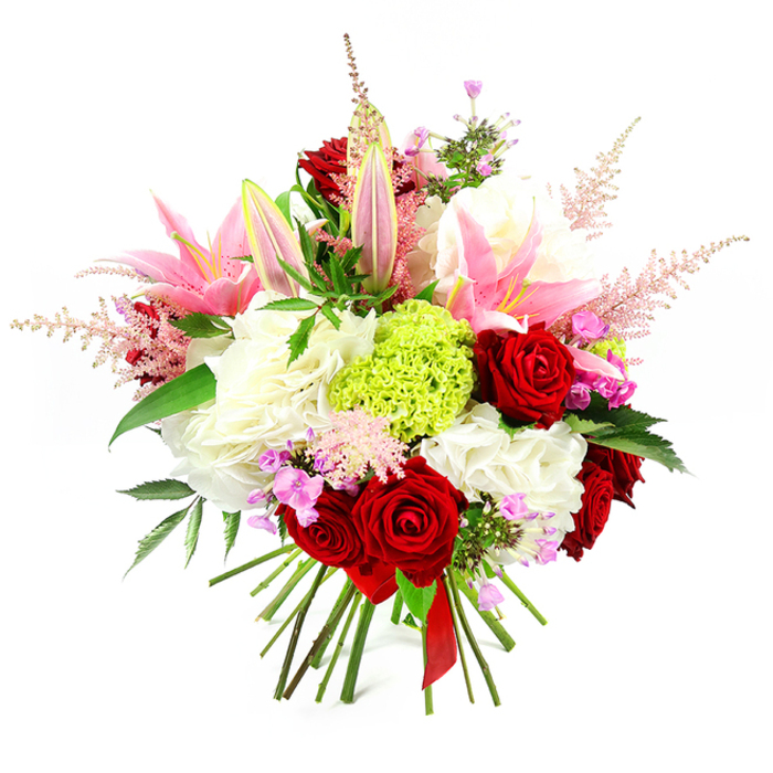 Against a bright white backdrop, this lavish bouquet appears almost sculptural, its carefully layered flowers forming a rounded, hand-tied arrangement immediately suggestive of celebration and care. Prominent among the blooms are several deep red roses, their petals velvety and rich, unfurling in tight spirals that create an intense, romantic focus. These roses are beautifully accompanied by pink lilies, which arc gracefully out from the centre; some are already open, displaying broad, gently curved petals and paler centres, while others remain in sleek buds, hinting at more colour and fragrance to come. Cradling these statement flowers is a substantial base of ruffled white blossoms, similar to hydrangeas or generous carnations, creating a plush, cloud-like foundation that lends body and luxury to the design. At the heart of the bouquet sits a distinctive lime green sphere of dianthus or carnation, its fine texture and vivid hue introducing a bold, contemporary accent reminiscent of fresh new growth in Mill Hill's leafy gardens. Soft, feathery plumes of pale pink astilbe emerge between the larger blooms, adding vertical interest and a sense of gentle movement, as though stirred by a light breeze on Mill Hill Broadway. Interspersed throughout the arrangement, tiny fuchsia and lavender filler flowers offer subtle pops of deeper colour, rewarding closer inspection and enriching the overall palette. Dark green foliage provides a lush backdrop, with glossy leaves slipping between the blossoms to give contrast, structure, and a natural garden feel. The stems are visible at the base, neatly trimmed and bound securely with a deep red satin ribbon, underscoring the bouquet's hand-tied craftsmanship. The composition feels both opulent and heartfelt, ideal for an anniversary dinner, a special birthday, or a striking gift delivered to a doorstep in Mill Hill.