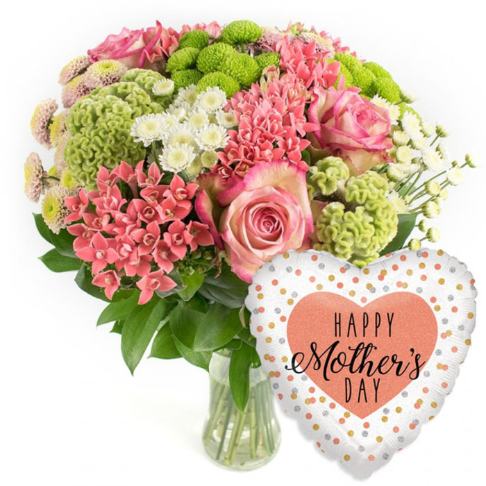 Photographed as if for an online catalogue for Flowers Mill Hill, this image showcases a luxurious Mother's Day bouquet paired with a heart-shaped balloon, all set against a pristine white background. The clear glass vase at the centre base reveals neatly trimmed green stems, gathered in cool water and supporting a beautifully balanced, rounded arrangement. Several prominent pink roses sit near the top and front, fully open and velvety, with petals that graduate from a softer inner blush to slightly richer pink edges, suggesting affection and gratitude. Nestled around these are generous clusters of coral-pink blooms, akin to kalanchoe, whose numerous tiny florets create a dense, jewel-like texture. Lime-green button chrysanthemums are carefully spaced throughout the bouquet, contributing bold, modern dots of colour and a unique, almost cushioned appearance that plays against the smoother rose petals. Small white daisy-style spray chrysanthemums and similar airy blooms thread in between, catching the light and giving the bouquet a fresh, uplifting quality. Deep green foliage frames the whole composition, peeking out at different levels and lending structure, much like the hedgerows and gardens that dot Mill Hill's leafy streets. To the lower right of the flowers, a glossy foil balloon in the shape of a heart rests against the invisible surface. It features a pale white-silver background scattered with irregular gold and soft peach-silver spots, like delicate confetti. Centered within is a larger peach heart that carries the inscription HAPPY Mother's DAY in black lettering, mixing playful bold capitals with a graceful script. The overall feeling is one of joyful celebration and tenderness, ideal for imagining a same-day delivery to a mum living near Mill Hill Broadway, along Bittacy Hill, or overlooking the green slopes toward Barnet.