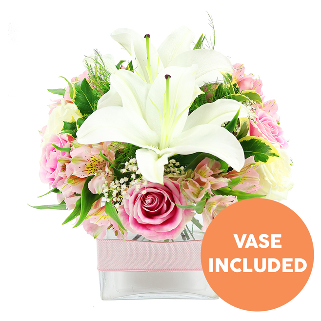 A beautifully composed bouquet in shades of white and pink is set against a bright white background, the kind of arrangement you might receive from Flowers Mill Hill on a special day. At the heart of the display, two impressive white Oriental lilies open fully, each flower forming a generous star with smooth, slightly curled petals and a cluster of dark reddish-brown stamens at its centre. Encircling these lilies is a full ring of blush and pastel pink roses, carefully spaced to show a progression from tight buds to fully opened blooms, their many layered petals overlapping in a soft, romantic swirl. Smaller heads of alstroemeria in pale pink and creamy white are threaded among the roses, their slim, pointed petals accented with light yellow flashes and fine speckling, giving the bouquet an intricate, almost hand-painted feel. Between these main flowers, airy stems of white gypsophila fill the gaps like tiny stars, adding a gentle, cloud-like haze that softens the overall silhouette. The blooms are supported and framed by a mix of lush green foliage: broad, glossy leaves, a few paler variegated pieces, and delicately feathery greens that evoke the natural textures of nearby woodlands around Mill Hill. All of the stems are gathered into a low, square clear glass vase, its transparency revealing the freshly cut stems and clean water inside, grounding the romantic colours in a contemporary container. Around the centre of the vase, a light pink satin ribbon is wrapped smoothly in a simple band, adding a hint of gift-wrapped elegance. In the bottom right corner of the image, a clear orange circular emblem with bold white capital letters reads VASE INCLUDED, making the composition appear ready to place straight onto a dining table or sideboard. The bouquet radiates thoughtful care and quiet joy, perfect for deliveries across Mill Hill and neighbouring streets to mark anniversaries, thank-yous, or gentle, meaningful occasions.