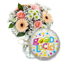 This image shows a thoughtfully composed 'Good Luck' gift set arranged on a crisp white background, the kind of uplifting package we prepare for neighbours across Mill Hill and surrounding streets. On the left a full bouquet nestles in a clear glass vase tied neatly with rustic twine; the blooms are a soft, harmonious mix of light pink roses and peach-toned Gerbera daisies whose darker centres give them weight and character. Fine sprays of white baby's breath (Gypsophila) thread gently through the composition like lace, while compact white-and-lime chrysanthemum clusters introduce a bright, modern accent. Dark green foliage provides contrast and a living structural frame so the pastel shades read clearly against the vase's glass.

Taking the foreground on the right, a round metallic foil balloon gleams with a silver-white sheen and a scattering of colourful celebratory motifs-stars, hearts and confetti-like dots in blue, pink, yellow and green. The bold phrase "Good Luck!" curves across the balloon in playful, multi-coloured letters; the two 'o's in "Good" are replaced by two sunny yellow smiley faces-one winking, the other pulling a playful face-bringing an immediate smile. The image feels like a personal send-off: under soft even lighting it radiates warmth and support, ideal for a student heading to exams, a friend starting a new job, or anyone in need of encouragement. I picture this being delivered from a local Mill Hill florist to homes around The Broadway or Mill Hill Park with care and quiet pride.