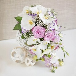 Captured as though already placed at the heart of an elegant Mill Hill reception, this floral arrangement combines contemporary design with classic romance. The flowers are gathered in a distinctive white spherical vase made from thin, interlaced strands, its open weave resembling a refined wicker ball or skein of decorative twine. It rests on a spotless white surface, joined by two smaller cream woven balls whose airy lattice echoes the vase and adds a playful sculptural touch. Rising from this stylish base are several radiant white Phalaenopsis orchids, their wide, velvety petals elegantly curved and each centred with a delicate splash of yellow and gold, lending an exotic, almost gallery-like quality. Woven closely around the orchids are lush roses in pastel pink and soft lavender, captured at varying stages of bloom so that some appear tight and promising, while others reveal glorious spirals of petals. Tucked between them, smaller white and lilac blossoms with purple-edged ruffles, reminiscent of Lisianthus, add fine detail, while wisps of white baby's breath drift through the arrangement, giving it a light, cloud-like softness. Trailing stems of light purple flowers cascade gently downward, suggesting fluid movement, as if the bouquet might be spilling quietly over the edge of a table set for dinner near Mill Hill Broadway. Polished green leaves and softer, fern-like foliage provide a fresh, leafy counterpoint to the pastel tones, framing the central blooms. The background is a pale, softly textured beige, blurred just enough to let every petal and woven strand stand out. Bathed in even, natural-style light, the entire design appears freshly arranged, ideal for illustrating wedding flowers, engagement parties, or sophisticated home entertaining in Mill Hill.