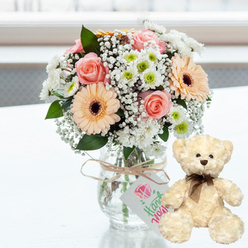 On a gleaming white tabletop, a carefully curated anniversary gift pairing is arranged in clear, gentle light, reminiscent of a calm morning in a Mill Hill home. The eye is first drawn to the central bouquet, a compact cluster of pastel blooms displayed in a simple clear glass jar vase. Inside, stems of blush pink roses, soft peach gerbera daisies with deep brown centres, and dainty white chrysanthemums tipped with lime green are gathered in a rounded shape that feels both elegant and approachable. Tiny white baby's breath blossoms float around the larger flowers like mist, filling the spaces and adding a sense of depth and airiness. Wrapped around the neck of the jar is a piece of natural twine, tied into a relaxed bow whose slightly frayed ends speak of artisanal care. From this bow hangs a small white tag printed with a pink heart or gemstone icon and slender green writing that suggests the gift has been thoughtfully crafted, not merely selected at random. To the right of the vase, a cream teddy bear is seated upright, facing slightly toward the viewer. Its fur is plush and textured, inviting the touch, while its dark brown eyes, matching nose, and neatly tied brown satin ribbon give it a look of gentle companionship. The softly blurred background, filled with bright white light, hints at a window view towards Mill Hill Park or the leafy streets off The Ridgeway, but remains neutral so nothing distracts from the main subjects. Altogether, this scene embodies quiet celebration and heartfelt sentiment, ideal for anniversaries, new baby welcomes, or comforting get-well wishes sent across Mill Hill and nearby areas.