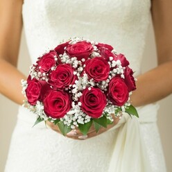 In the heart of Mill Hill, just moments from the tree-lined avenues and village greens, this classic bridal bouquet takes centre stage. A lush dome of deep red roses, immaculately arranged, draws the eye-each blossom is full and luxuriously layered, embodying both passion and poise. Baby's breath, light as morning mist, weaves between the roses in soft white clusters, echoing the purity and delicacy of a wedding day. At the bouquet's base, sprigs of vibrant green offer a fresh, organic accent, reminiscent of Mill Hill's open spaces. The bride's hands, refined and carefully manicured, wrap tenderly around the stems, cradling the artful composition with a sense of devotion. Her wedding dress-textured with intricate lacework and a gentle draped sash-adds another layer of elegance, reflecting the neighbourhood's timeless charm. The neutral, blurred background ensures every eye is drawn to the bold reds, snowy whites, and natural detail of the arrangement. Perfect for discussions of bridal flowers, wedding inspiration, or London romance, this bouquet is a testament to Mill Hill's floral expertise and the artistry of Flowers Mill Hill.