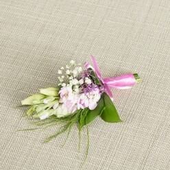 A refined boutonniere or petite corsage lies perfectly composed on a pale, woven linen backdrop, the neutral beige threads creating a warm, understated contrast to the flowers' soft colours. The focal point is a small cluster of blush pink, multi-petalled blooms, their ruffled petals layered densely so that each flower looks like a miniature garden in itself. The pinks move gently from almost-white at the edges to a more saturated rose tone at the heart, giving the arrangement a subtle depth. Nestled among these are fine sprays of white baby's breath, each tiny bloom like a dot of lace that lightens and softens the whole piece. Slim stems of lavender or deep purple florets peek through the pastel blossoms, adding a hint of richness that keeps the palette from feeling too sweet, more like an early evening sky over Mill Hill than a simple pink-and-white mix. On one side, a fan of closed creamy white buds tipped with pale green leans outward, evoking young freesia or similar delicate blooms just waiting to unfold. Lush, broad green leaves support the composition, while fine, wispy strands of fern-like greenery stretch out from beneath, suggesting movement and a slightly natural, garden-grown character, as though gathered from a carefully tended border near Mill Hill East. The stems are wrapped with precision in a bright fuchsia satin ribbon, tied into an elegant bow with soft tails that rest lightly on the fabric. The lighting is gentle and even, highlighting each texture and curve, making this accessory feel perfect for a local Mill Hill wedding, engagement photoshoot, or prom night stroll along the quiet residential streets.