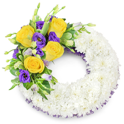 There is a calm craftsmanship in this circular funeral wreath: a continuous ring of tightly packed white spray chrysanthemums forms a plush, almost pillow-like base whose small, overlapping petals scatter soft highlights and shadows. This white halo, dense and immaculate, suggests purity and a steady quiet. From the upper left a sculpted spray arcs outward-a cluster of warm yellow roses, their petals unfolding like tiny suns, paired with rich purple lisianthus that present satiny, ruffled cups and a few pale lavender-to-green buds that add life and promise. Glossy green foliage-rounded salal-like leaves, some with faint variegation, and slender fern fronds-threads through the bloom cluster, supplying texture and a cool, natural frame. A slim band of dark purple mesh or crinoline peeks from under the chrysanthemum edge, a subtle finishing detail that links the accent flowers back to the wreath's core. The colour play is deliberate: the white speaks of peace, yellow of friendship and warmth, purple of dignity and solace. Light falls softly across the arrangement, accentuating petal edges and the delicate scent you might imagine from roses and chrysanthemums. For families in Mill Hill Village or those organising services near Mill Hill Broadway, this composition reads as a carefully considered tribute-refined, respectful and handcrafted with the sensitivity of an experienced local florist preparing farewell flowers for a close-knit community.