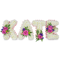 This upright floral name tribute spells KATE in four sculpted letters, each carefully formed from thousands of small, white chrysanthemums to produce a dense, velvety surface that reads pure and serene against the bright white background. A soft pink satin ribbon outlines every letter, the sheen catching morning or chapel light and adding a blush border that defines the shapes without overpowering them. Each letter is embellished with vivid accents: large, velvety fuchsia-pink roses paired with delicate pastel pink spray roses, and fine sprays of deep violet or blue lisianthus or statice, which sit like thoughtful brushstrokes against the white chrysanthemum canvas. Varied foliage-glossy eucalyptus and delicate fern fronds-creates a natural frame, introducing texture, movement, and a cool green note to the composition. You can picture the placements: the K bears a pronounced floral cluster along its diagonal stroke; the A is brightened near its lower right curve; the T holds a supportive cluster at its base; while the E features two measured groupings on its middle bar and bottom arm. The overall effect is solemn yet tender, with a faint floral perfume and the subtle rustle of ribbon that comforts more than it declares. Designed as a sympathy or memorial piece, it makes a dignified statement for a funeral or celebration of life in Mill Hill, suitable for delivery around Mill Hill Broadway and local chapels, where its craftsmanship and quiet emotion will be deeply felt.