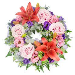 Photographed from above on a bright white background, this sympathy wreath feels both abundant and thoughtfully restrained, ideal for a quiet farewell in Mill Hill. The design forms a perfect circle, its continuity emphasised by two striking reddish-orange Asiatic lilies set across from one another. Their broad petals, dusted with pollen and open to reveal strong stamens, bring warmth and a sense of courage to the tribute. Between them, generous groupings of blush pink roses unfold layer upon layer of velvety petals, evoking gentleness and a deep, caring love. Woven among the roses are lisianthus blossoms in pale pink and sumptuous dark purple, their softly ruffled edges creating a fabric-like texture that softens the stronger lines of the lilies. Fine, cloud-like sprays of lavender-blue statice peek out from the arrangement, introducing a light, airy quality and subtle fragrance. The entire circle rests on a bed of lush greenery, combining rounded, silvery eucalyptus leaves, delicate fern fronds, and darker, more structured foliage. Spiky blue eryngium thistles punctuate the greens with cool, architectural bursts, offering visual interest without overpowering the rest of the flowers. Several closed lily buds are nestled discreetly within the foliage, hinting at new blooms and the enduring nature of memory. Bright green, folded leaves, likely Ti or aspidistra, are wrapped artfully around the bases of the lilies, lending a modern, polished finish. This wreath would feel at home in a chapel near Mill Hill East station or at a family service off Lawrence Street, offering a dignified expression of sympathy and quiet support.