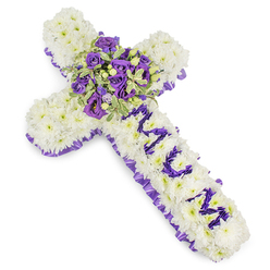 This cross-shaped sympathy tribute is tailored with intentional care, the kind of work we prepare for services across Mill Hill and surrounding Barnet streets. The main field of the cross is completely covered in compact white chrysanthemum florets, their downy texture creating a serene, cloud-like base that absorbs light softly on the white backdrop. A ruffled band of deep purple satin is woven around the perimeter, adding a dignified frame that lifts the cooler tones. The vertical arm carries the personalized message MUM, each letter formed from smaller purple blooms - likely eustoma or spray chrysanthemums - placed densely to read clearly against the white. The centre focal spray is a luxuriant cluster of deep violet and lavender roses with pale lilac lisianthus, accented by variegated foliage whose pale edges catch light and give the bouquet depth; small green berries add a subtle, natural punctuation. The arrangement breathes a restrained elegance: the visual weight is balanced, the texture moves from pillowy white to velvety purples, and there's a soft, floral trace in the air that comforts rather than overwhelms. Prepared by Flowers Mill Hill for families in Mill Hill Village, Mill Hill Broadway and the Ridgeway, this piece is suited for wakes, graveside services, or a chapel tribute - a composed, heartfelt expression of love and remembrance.