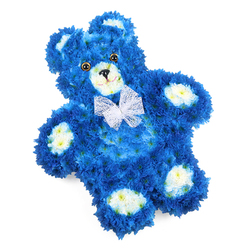 Against a crisp white backdrop, a tender teddy bear fashioned entirely from flowers seems to float, the kind of piece that might rest on an easel in a Mill Hill funeral chapel or by a memory table in a family home near Mill Hill Park. The bear is composed of densely packed royal blue blooms, most likely small chrysanthemum heads, meticulously arranged so that they form a seamless, cushion-like coat of petals. This rich blue gives the figure a sense of depth, with darker tones appearing in the curves of the limbs and lighter highlights along the top of the head and ears. Soft clusters of creamy white blossoms, each with a subtle wash of pale yellow at the centre, define the snout, the inner ears, and the four oval paw pads, gently interrupting the blue and guiding the eye to the bear's key features. The face is simple yet expressive: two round, polished eyes in deep brown or amber sit evenly on the blue background, above a small, black, inverted triangle nose. Just beneath, a fine curved black line suggests a faint smile, giving the bear a comforting, almost guardian-like presence. Around its neck, a delicate bow of white mesh or organza is tied in neat loops, its sheerness adding a light, innocent flourish. The whole figure is slightly rotated, which softens the composition and makes the bear appear to be quietly resting. Created with the care of an experienced florist, this floral teddy feels particularly fitting as a tribute for a baby boy, a child, or a young-at-heart loved one in Mill Hill, where its blend of vivid blue and gentle creams speaks of remembrance, purity, and deep affection.