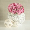 A soft, romantic still life shows a rounded bouquet of pink roses and white baby's breath displayed on a clean white surface, styled with simple decorative orbs. At the centre of the image sits a compact dome of around fifteen to twenty soft pink roses, each bloom cupped closely to the next so the petals almost touch, forming a perfect sphere of colour. The roses are two-toned: their centres glow with a gentle blush that gradually deepens towards the outer edges into a richer, almost fuchsia pink, like the evening sky over Mill Hill Park just before sunset. Threaded delicately between the roses are fine sprays of white baby's breath, their tiny cloud-like blossoms softening the structure of the bouquet and adding an airy, bridal lightness. The flowers are nestled in a spherical white vase made from intricately woven material that resembles painted wicker or rattan, with an open criss-cross pattern. Light spills through these gaps, casting faint, lacy shadows on the surface beneath, hinting at handcrafted detail and a slightly rustic charm that would suit many Mill Hill homes. To the left of the vase, two decorative woven spheres in pale natural beige echo the texture of the container, one larger and one smaller, resting casually on the tabletop. They contribute a modern, organic accent, as if chosen from a local interiors boutique off Mill Hill Broadway. The background is a soft, textured wall in warm beige, gently lit with bright but diffused light that eliminates harsh shadows. Overall, the composition feels calm, elegant, and lovingly arranged, suggesting a peaceful corner bouquet ideal for birthdays, anniversaries, or simply bringing a touch of romance into a North London living room.