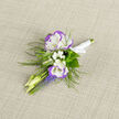 This boutonniere is a miniature study in gentle colour and texture: two lisianthus blooms, their petals white at the heart and edged with a cool violet blush, anchor the arrangement with quiet elegance. Around them, sprays of tiny white baby's breath create an airy cushion, while slim pale buds and short clusters of grape hyacinth add a lively vertical rhythm in clear blue-violet. Feathery fern fronds weave through the stems, offering a woodland softness that contrasts with the clean fold of the white satin ribbon knotted at the base. The fabric of the ribbon and the textured linen beneath give a tactile counterpoint to the velvety lisianthus petals; the boutonniere smells faintly of green freshness rather than heavy floral perfume, an understated aroma that suits close-up moments-a groom pinning it on at the altar, a portrait session on a cool spring afternoon. As a local Mill Hill florist, I assemble pieces like this for ceremonies and small celebrations across Mill Hill Broadway and the quieter lanes near The Ridgeway; Flowers Mill Hill prepares each pinned piece to sit comfortably on a jacket lapel, balanced to catch the light and draw the eye without shouting. It's a compact, carefully composed accent that speaks of intention, memory, and the tender details that make a day feel handcrafted and personal.