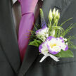 Nestled against the deep charcoal weave of a well-cut suit, this boutonnière radiates composed elegance and considered artistry, the kind we craft at Flowers Mill Hill for weddings and special occasions across Mill Hill Broadway and Mill Hill Village. At its centre sits a white lisianthus with petals edged in vibrant purple, each ruffle catching light like a natural highlight that frames the bloom. Slim blades of fresh green foliage cradle the flower, their sheen and movement giving the arrangement an organic, lively silhouette, while tiny sprays of gypsophila add a whisper of innocence and lift. An upright stem of indigo veronica introduces a refined vertical line; nearby, three slender lisianthus buds promise gentle unfolding - a quiet narrative of hope and newness. The entire cluster is secured with a crisp white ribbon whose smooth texture complements the satiny petals and the adjacent magenta tie, producing a sophisticated contrast against charcoal fabric. A faint, verdant fragrance and the subtle rustle of leaves heighten the sensory experience, comforting through long ceremonies and celebratory evenings. Picture this pinned at a reception near The Ridgeway or carried from a photoshoot around the green spaces by Mill Hill East: it's both modern and timeless, a small emblem of care that enhances portraits and lingers in memory. This boutonnière embodies skilled floristry, local familiarity, and an attention to detail that feels both personal and perfectly suited to important days.