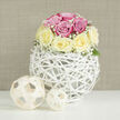 This image captures an elegant, modern rose arrangement that feels perfectly at home in a stylish Mill Hill interior, perhaps near the calm streets off Mill Hill Broadway. Filling the centre of the frame is a rounded bouquet of fresh roses, meticulously shaped into a low, spherical dome. The flowers alternate between soft romantic pink and a buttery, pale yellow, their petals lush and velvety, layered in tight spirals that invite the eye to linger. Fine sprays of white baby's breath weave between the roses, creating a light, misty fringe and adding an impression of lace around the bouquet's edge. A few muted green leaves nestle at the base, gently grounding the pastel tones without breaking the overall softness. The bouquet is cradled in a distinctive white orb-shaped vase made from thin, interlaced rattan-like strands, forming an open lattice that feels both sculptural and airy. The structure's curved lines and gaps let the light pass through, casting intricate shadows onto the pristine white tabletop. Flanking the arrangement are two decorative wooden spheres in a natural, light beige hue, their woven construction mirroring the vase's pattern and adding texture and balance to the scene. The background is a warm, neutral beige with a faint linen-like horizontal texture, keeping the focus firmly on the flowers. Soft, even lighting creates minimal shadows and a sense of calm clarity, as though this piece has been placed in a bright Mill Hill home just before guests arrive. The composition evokes understated luxury and heartfelt intention, ideal for celebrating an anniversary, saying thank you, or sending a gentle congratulations with local flower delivery from flowersmillhill.info.