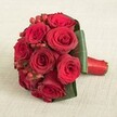 Arranged for a moment of love and significance in the heart of Mill Hill, this abundant bridal bouquet features eight to ten impeccably formed crimson roses, their petals plush and glowing with a deep, passionate red. Interspersed among the roses, vibrant hypericum berries gleam like tiny festive ornaments, bringing youthful texture and joyful detail. The bouquet's form is given definition by broad, glossy green leaves which encircle the blooms, providing both strength and an elegant visual frame. The stems are expertly gathered, bound by a radiant red satin ribbon that forms a smooth handle-functional, yet as sophisticated as a Mill Hill formal gathering. Set upon a neutral, slightly textured linen, the bouquet is bathed in even daylight that reveals every intricacy, from the gentle undulation of the leaves to the subtle gradations on each petal. More than a floral arrangement, it is an invitation to celebrate, to commemorate love and union in NW7, and to trust in the careful touch and local understanding of Mill Hill's floristry professionals. The overall mood-romantic, uplifting, and superbly crafted-invites admiration and promises memories cherished for years to come.
