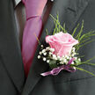 Pinned with care to a deep charcoal jacket, this blush-pink rose boutonniere radiates a gentle, timeless charm, as if it were made for a romantic ceremony overlooking the green spaces of Mill Hill. The rose itself is the heart of the piece: its petals are soft and velvety, curling outward in a near-perfect rosette, the color shifting from pale, almost porcelain pink at the center to a deeper, warmer blush toward the edges. Draped around this central bloom are airy tufts of white gypsophila, or baby's breath, their many tiny flowers clustered so closely they resemble a cloud drifting around the rose, lightening the overall look and adding a whisper of texture. Delicate sprays of fresh greenery peek from behind, their fine lines extending just beyond the edges of the flower to give the boutonniere a graceful silhouette that sits naturally against the structured suit fabric. The stems are tightly gathered and wrapped in a narrow satin ribbon, likely in a lilac or soft mauve tone, smooth and secure, its subtle sheen catching the light without drawing focus from the rose. The lighting is gentle and even, suggesting a preparation room or florist's studio near Mill Hill Broadway, where the petals are carefully checked and adjusted before the piece is delivered for a wedding or prom. You can almost imagine the faint fragrance of the rose, clean and sweet, lending a final touch of sensory richness. This design feels intentional and heartfelt, a small but meaningful floral accent that complements anything from a traditional church ceremony by Mill Hill's quiet streets to a modern celebration in a local hotel.