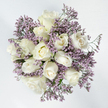 The photograph shows a circular bouquet composed of white roses and lavender limonium, viewed from directly above against a pristine white background that enhances its gentle, contemplative atmosphere. At the centre, around a dozen white roses form a dense, rounded cluster. Each bloom varies slightly: some are tightly furled, with outer petals hugging close to the centre, while others are more open, revealing their layered inner whorls with a soft, spiralling motion. The roses are a creamy, elegant white rather than a harsh bright tone, with the slightest suggestion of ivory and pale butter at the core that adds depth and warmth. Around and between these central blooms, airy sprays of lavender limonium extend outward, their many tiny flowers creating a delicate, mist-like halo of lilac and soft purple. This filler flower gives the bouquet an ethereal, floating quality, offsetting the solid, velvety presence of the roses. Tiny hints of green from stems and the most minimal foliage peek from beneath, grounding the arrangement in nature without disturbing the dominant white and lavender theme. The composition feels serene and balanced, making it an especially fitting design for sympathy flowers or quiet memorial arrangements. In Mill Hill, a bouquet like this might be chosen for a thoughtful delivery to a home near Mill Hill Park or a flat overlooking the gentle slopes of the area, where its calm beauty would offer solace, convey sincere condolences, and bring a touch of peaceful grace to a room in a time of loss.