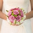 Bathed in gentle morning light, a Mill Hill bride stands poised, her bouquet glowing with quiet excitement and love. In her hands rests a symphony of colour: roses in rich lavender and dusty mauve, their outer petals tinged with fuchsia, are embraced by a lush collar of lime-green button chrysanthemums, whose sculpted orbs bring lively texture and playful contrast. Framing the floral heart, a halo of snowy white, possibly formed by fragrant freesias or dainty clusters of spray roses, adds a touch of classic refinement and fresh fragrance to the composition. Wisps of green buds peek through, gently threading the arrangement together. The bride's soft ivory gown, adorned with detailed lacework reminiscent of the architectural charm of Mill Hill Broadway, serves as an elegant canvas, while her manicured nails-rosy pink with a sparkle on her ring finger-echo the celebration's joy. This bespoke arrangement, crafted for local couples marrying near Mill Hill Park or in the timeless surrounds of north London's many chapels, is more than a bouquet: it's a sensorial keepsake of artistry and devotion, capturing the essence of weddings in the heart of Mill Hill.