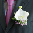 This boutonniere features a single, radiant white orchid, pinned with precision to a charcoal suit and glowing softly against the deep fabric, like the first light over the rooftops of Mill Hill. The orchid's petals are gently cupped and wonderfully smooth, almost porcelain-like in their sheen, with a hint of creamy depth near the outer edges that adds warmth to the cool white. Embracing the central bloom, pale green and silvery leaves create a graceful frame, while threads of lavender flowers and tiny white buds weave through the greenery, offering delicate detail that only reveals itself up close-much like the quiet charm of the side streets around Mill Hill Broadway. The stems are bound in a slim lavender-violet ribbon, tied with careful, practiced hands, its soft shimmer echoing the lilac tie beneath and tying the whole look together with understated sophistication. The composition feels both artful and effortless, the mark of an experienced local florist used to dressing grooms for intimate ceremonies near Mill Hill Synagogue or stylish receptions along The Ridgeway. As you picture this orchid resting on the lapel, you can sense the anticipation in the air: the last adjustments in the mirror, the reassuring touch on the shoulder from a best man, and the gentle hum of family and friends gathering nearby. This is more than a simple floral accessory; it is a fresh, living symbol of celebration and tenderness, ready to accompany its wearer from the first greeting to the final dance of a perfect day in Mill Hill.