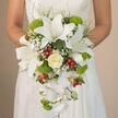 In this intimate bridal portrait, the camera lingers on a cascading bouquet that seems to flow like water against the subtle off-white lace of a wedding gown. The design centres on several striking white oriental lilies, each blossom wide open and confident, their broad petals smooth and slightly curved, marked with tiny reddish-brown speckles that draw the eye towards the pollen-dusted stamens. Just beneath them, a creamy ivory rose sits like a quiet promise, its many layered petals tightly cupped and softly glowing. Around these main blooms, little clouds of white baby's breath drift between the flowers, creating gentle, airy pockets of space so the eye can rest. From the lower edge of the bouquet, elegant white Phalaenopsis orchids cascade in loose strands, their slender petals and pale yellow throats adding movement and a sense of gentle luxury. Bright lime-green pompon blooms or Green Trick dianthus appear in rounded, fuzzy mounds, giving a playful, tactile element that contrasts beautifully with the smooth lilies. Scattered among these textures, glossy red hypericum berries add sparkling accents, each small sphere catching the light against the darker green foliage. The bouquet fills the frame with its teardrop shape, tapering gracefully downwards in a way that would sway softly as the bride walks along a quiet Mill Hill lane or steps into a local venue near Mill Hill Broadway. The overall effect is polished yet organic, a refined floral composition that feels right at home in this leafy North London neighbourhood, perfect for a couple seeking understated drama on their wedding day.
