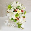 An elegant, cascading floral arrangement appears in the centre of the image, poised on a white tabletop that reflects a soft glow, with a neutral beige textured backdrop lending warmth and calm. The flowers are gathered in a round, white woven basket, whose subtle wicker pattern brings a handcrafted charm that feels right at home in Mill Hill's village-like atmosphere. Rising from the basket, several large white lilies unfurl their trumpet-shaped petals, revealing speckled throats and golden stamens lightly dusted with pollen, suggesting a gently sweet, classic fragrance. Slightly above the central cluster, a single creamy, pale-yellow rose sits in partial bud, its tightly layered petals adding a tender focal point and a hint of warmth amid the cooler whites. The entire arrangement is enveloped in lush greenery: broader glossy leaves form a base, while slender, feathery fronds and wispy grasses extend outward, adding softness and movement. Tiny white flowers, reminiscent of baby's breath, are scattered throughout, giving the bouquet a delicate, cloud-like texture. On the right side, a cascade of white Phalaenopsis orchids tumbles gracefully down from the main body of the design, their smooth petals and gentle curves creating a waterfall effect that would look striking along the edge of a bridal table. Nestled among the foliage are bright green pom-pom blooms, their spherical, almost mossy texture adding contemporary flair, while clusters of deep ruby-red berries tuck into the design, offering glossy, jewel-like accents. To the left, two white decorative spheres with an open lattice construction rest on the table, echoing the basket's weaving and balancing the composition. The overall effect is one of serene luxury and celebration, ideal for wedding receptions, anniversaries, or formal gatherings throughout Mill Hill, with Flowers Mill Hill ready to recreate this refined look for local events.