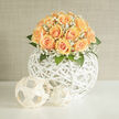 This hand-crafted composition marries soft peach roses with frothy clouds of white gypsophila, forming a rounded, tactile bouquet that feels both modern and timeless. Each rose unfurls in concentric spirals, a buttery dance of petals that shifts from pale cream to apricot, while the baby's breath threads through like silk, catching highlights and adding an ethereal hush. The flowers are arranged atop a sculptural white woven sphere - an openwork rattan orb whose criss-crossing lines create pockets of light and shadow, lifting the blooms with an airy, architectural grace. Green foliage peeks from beneath, grounding the palette and adding a faint green, resinous scent to the sweeter rose perfume.
The scene is softly lit, with warm daylight skimming each petal and giving the whole arrangement a faint golden halo. Perfect for a wedding, a milestone celebration, or as a refined centrepiece in a Mill Hill home, the bouquet evokes the quiet joy of new beginnings. I imagine it arriving by hand to a flat near Mill Hill Broadway or being chosen by a couple exchanging vows close to local parks - a design that reads beautifully in both contemporary apartments and village-style interiors. Prepared with the attentive eye of a local florist, this piece balances texture, tone, and form, and is ideal for customers seeking a graceful statement that feels personal and distinctly of the neighbourhood.