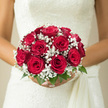 This image shows a rounded bridal bouquet composed of lush red roses and a halo of white baby's breath, held close against a softly textured ivory gown. The roses form the central focus, their blooms packed tightly together to create a full, domed shape, each flower a deep, romantic crimson that suggests warmth, loyalty, and enduring affection. Petals are smooth and velvety, curling outwards from dark, shadowed centres into brighter red edges, giving the bouquet a sense of depth and richness. Edging the roses, baby's breath provides a delicate contrast, its tiny white flowers clustered densely enough to look like a gentle mist, lightening the visual weight of the arrangement. Just beneath, a collar of fresh green foliage subtly supports the design, evoking the manicured greenery and peaceful gardens that characterise Mill Hill's quieter corners. The bouquet appears expertly arranged by a local florist, the stems likely bound neatly and concealed so that only the blooms and their textures are emphasised. Soft, even lighting brings out the gentle sheen on each petal and the intricate details of the gypsophila, while the neutral tone of the dress behind ensures the colours remain the clear stars of the composition. The overall impression is of a classic, London wedding moment: perhaps the bride stands outside a registry office near Mill Hill Broadway or pauses in a nearby park for portraits, bouquet held slightly forward as a focal point of the day. One can almost sense the quiet murmur of guests and the subtle perfume of roses, creating a tender atmosphere as two lives begin a new chapter together.